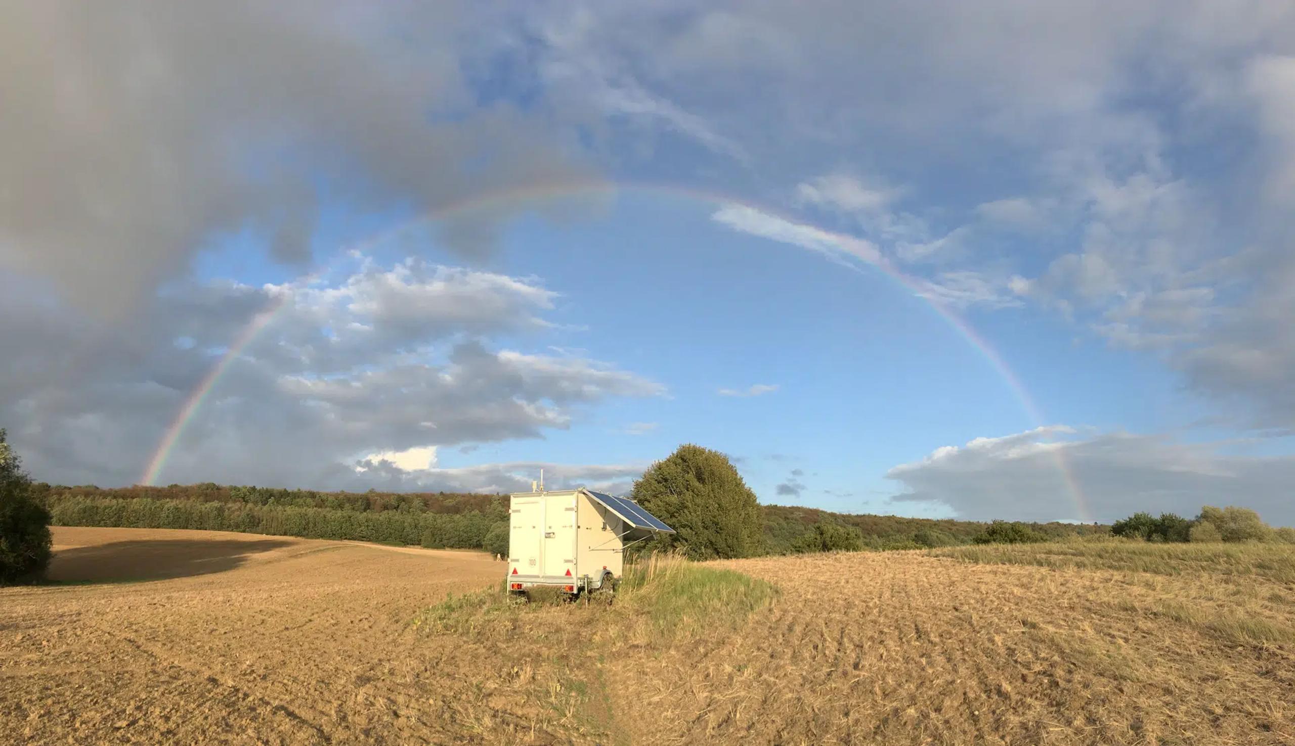 Messtrailer mit aufgestelltem Solarmodul auf einem abgeernteten Feld, darüber ein Regenbogen vor bewölktem Himmel.