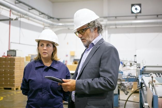 Une responsable de production et un collègue casqués consultent des informations sur une tablette dans l’atelier