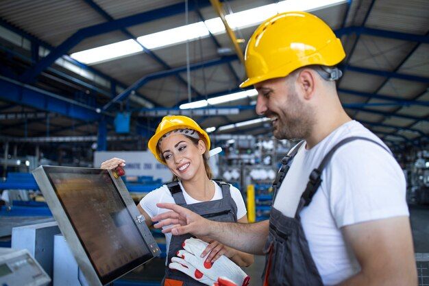 A smiling woman and a man, both in yellow hard hats and work overalls, discuss information on a control screen inside a factory hall