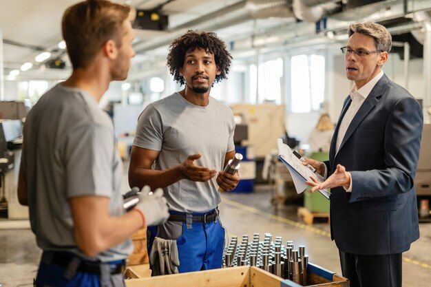 A manager holding a clipboard discusses a production process with two workers beside a crate of machined metal parts