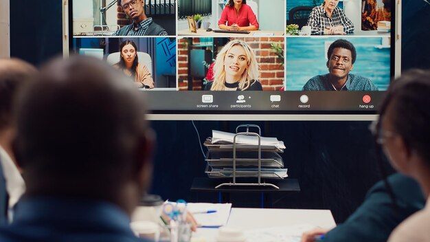A team in a meeting room watches a wall screen showing a video call with several remote colleagues