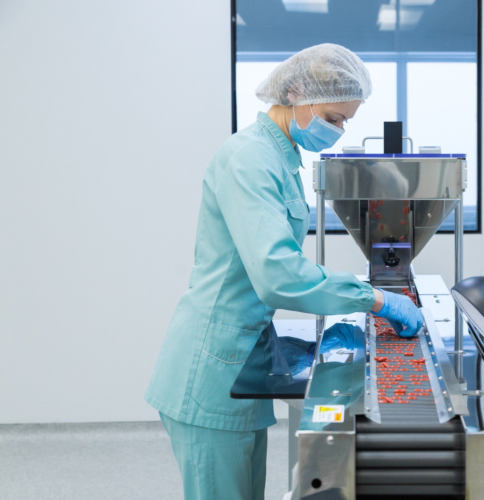 An operator in a cleanroom suit, hairnet and mask checks tablets on a production line