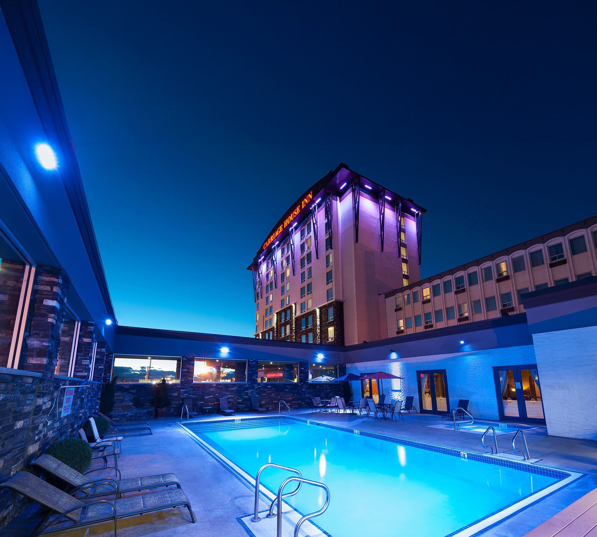 The exterior of Carriage House Hotel at night, with a focus on the patio and outdoor pool, which is surrounded by lounge chairs.