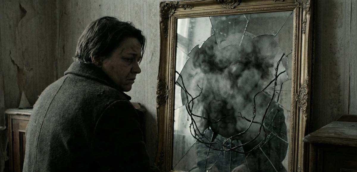 Uma mulher de meia-idade, com cabelos curtos e grisalhos e vestindo um casaco de lã grosso e cinza, é fotografada de perfil, de costas para a câmera, olhando para a esquerda. Seu rosto, visível de perfil, expressa tristeza e cansaço. Ela está sentada em frente a um espelho antigo e grande, com uma moldura de madeira entalhada e dourada. O espelho está quebrado em grandes pedaços. O reflexo não mostra a mulher, mas sim uma massa escura e caótica de fumaça cinza e galhos retorcidos, como se fosse uma tempestade interior. A sala onde a cena ocorre é velha e mal iluminada, com papel de parede descascado, móveis de madeira desgastados e uma atmosfera sombria. As cores são suaves e dessaturadas, em tons de cinza, marrom e dourado envelhecido.