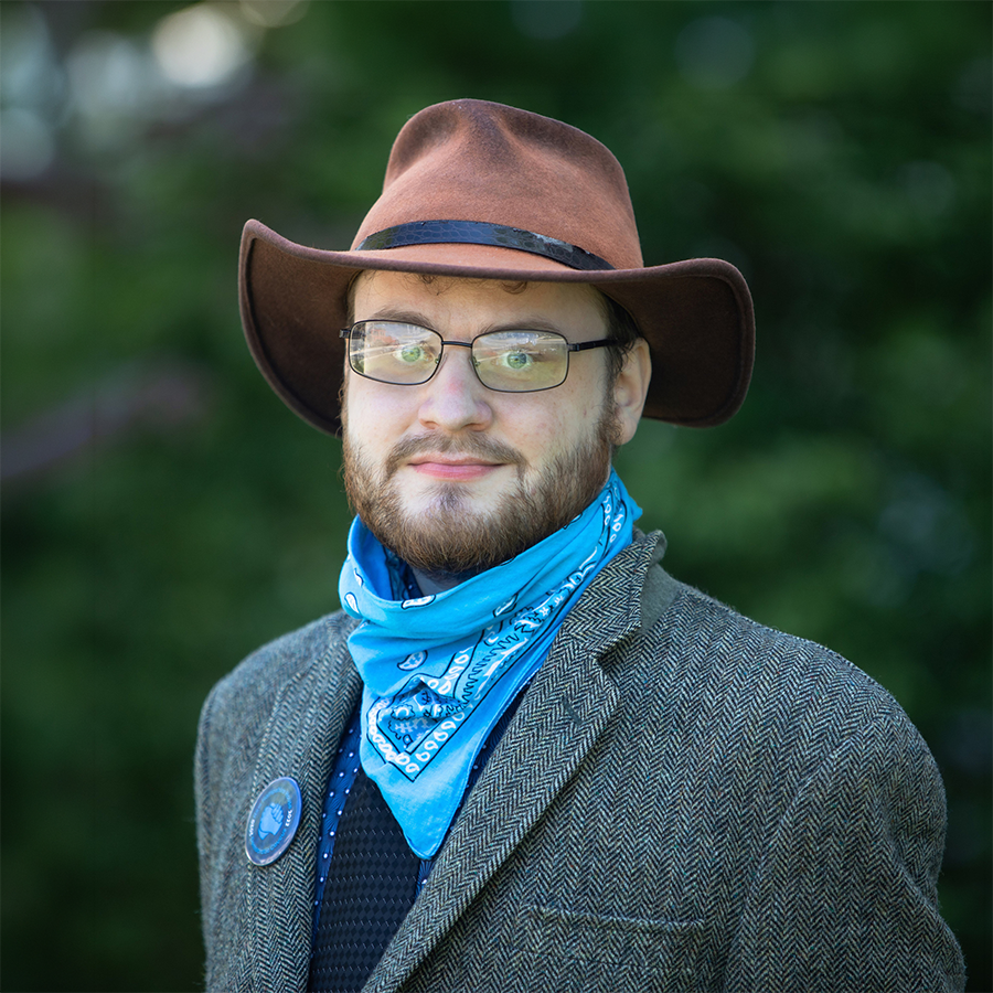 Headshot of Eric Gallager smiling slightly, wearing a brown hat, glasses, a patterned blue bandana, and a tweed jacket against a background of green trees
