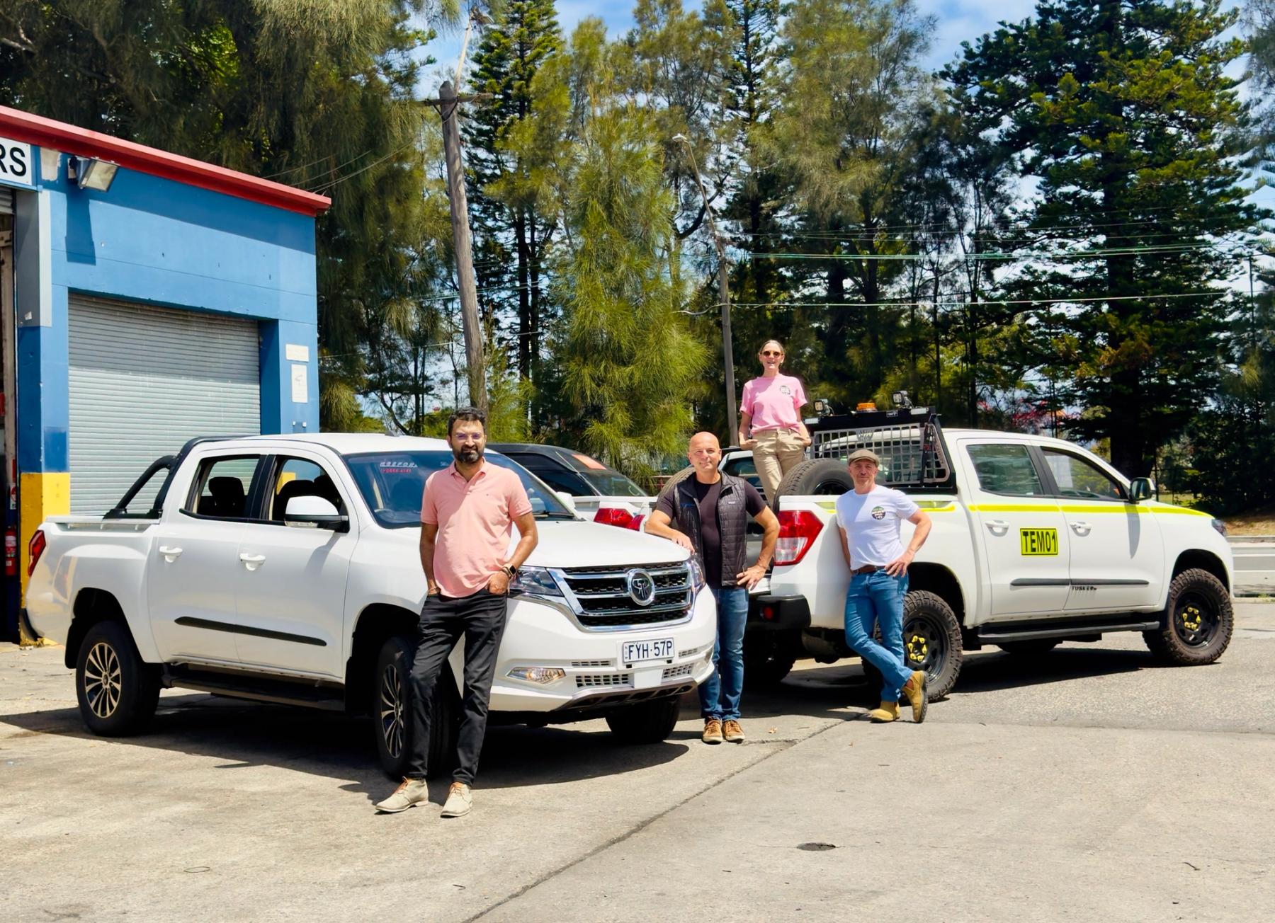 Four people pose with a Tembo Tusker electric dual-cab ute and another white dual-cab ute outside a workshop