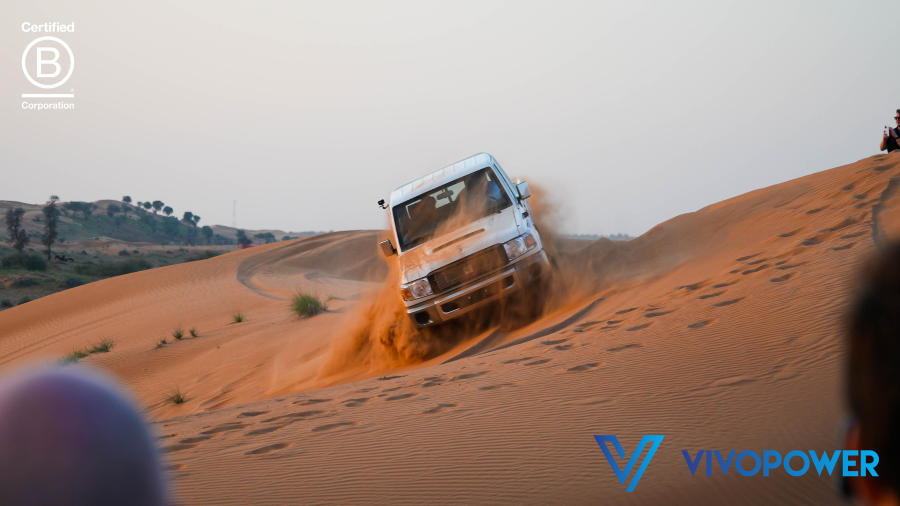 White electric Land Cruiser single-cab ploughing through a red sand dune in a spray of sand, spectators in foreground