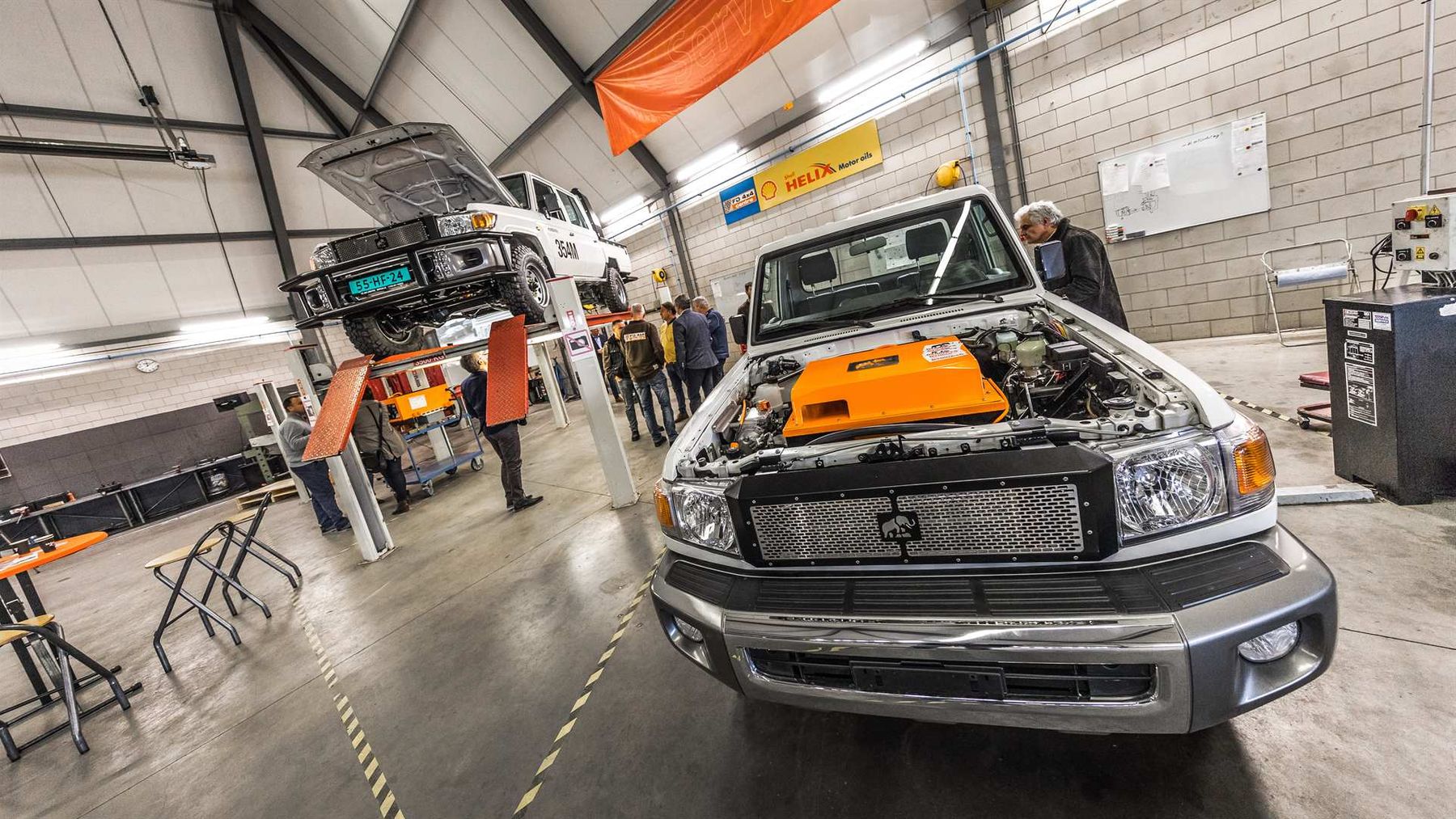 Electric Toyota Land Cruiser 70 conversion with bonnet open in a workshop, as visitors gather by another on a hoist