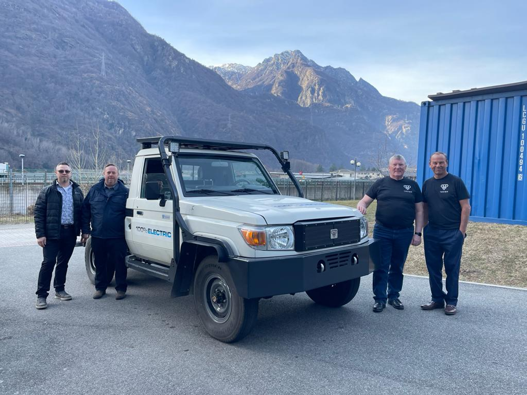 Two Tembo staff and two guests beside a 100% electric Land Cruiser 70 single-cab ute, with mountains behind