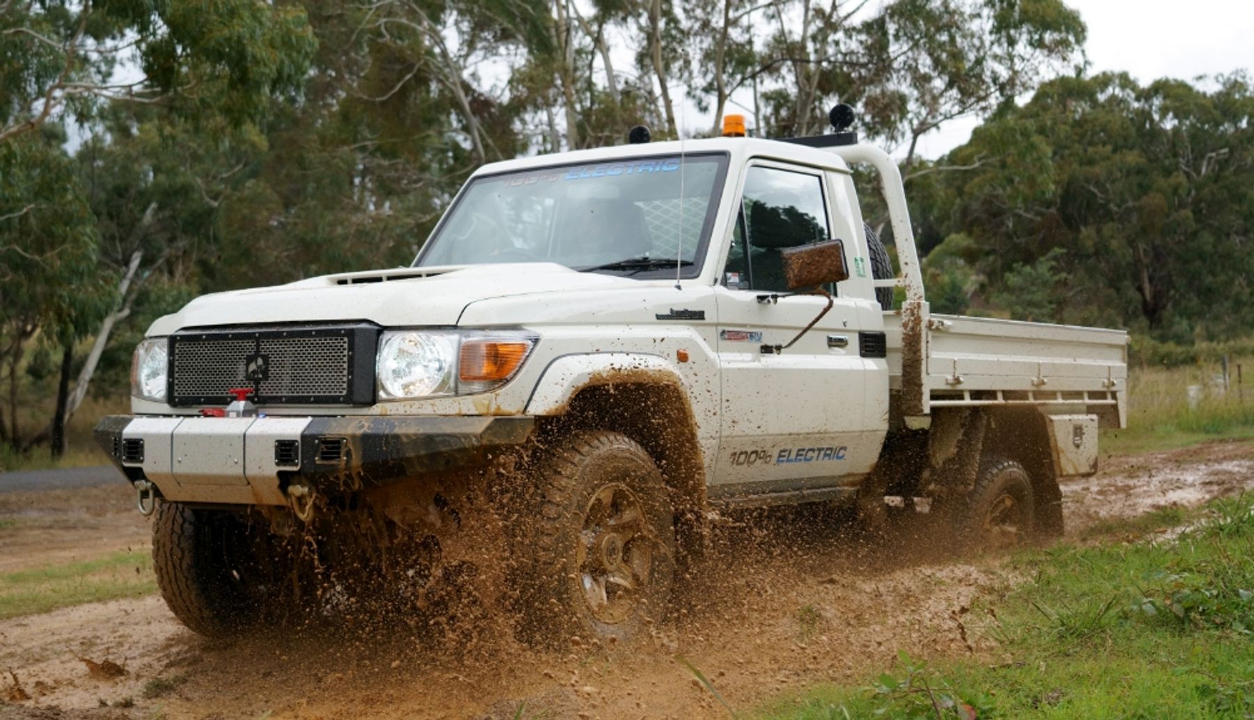 White electric Land Cruiser 70 single-cab tray ute marked 100% electric splashing through mud on a bush track