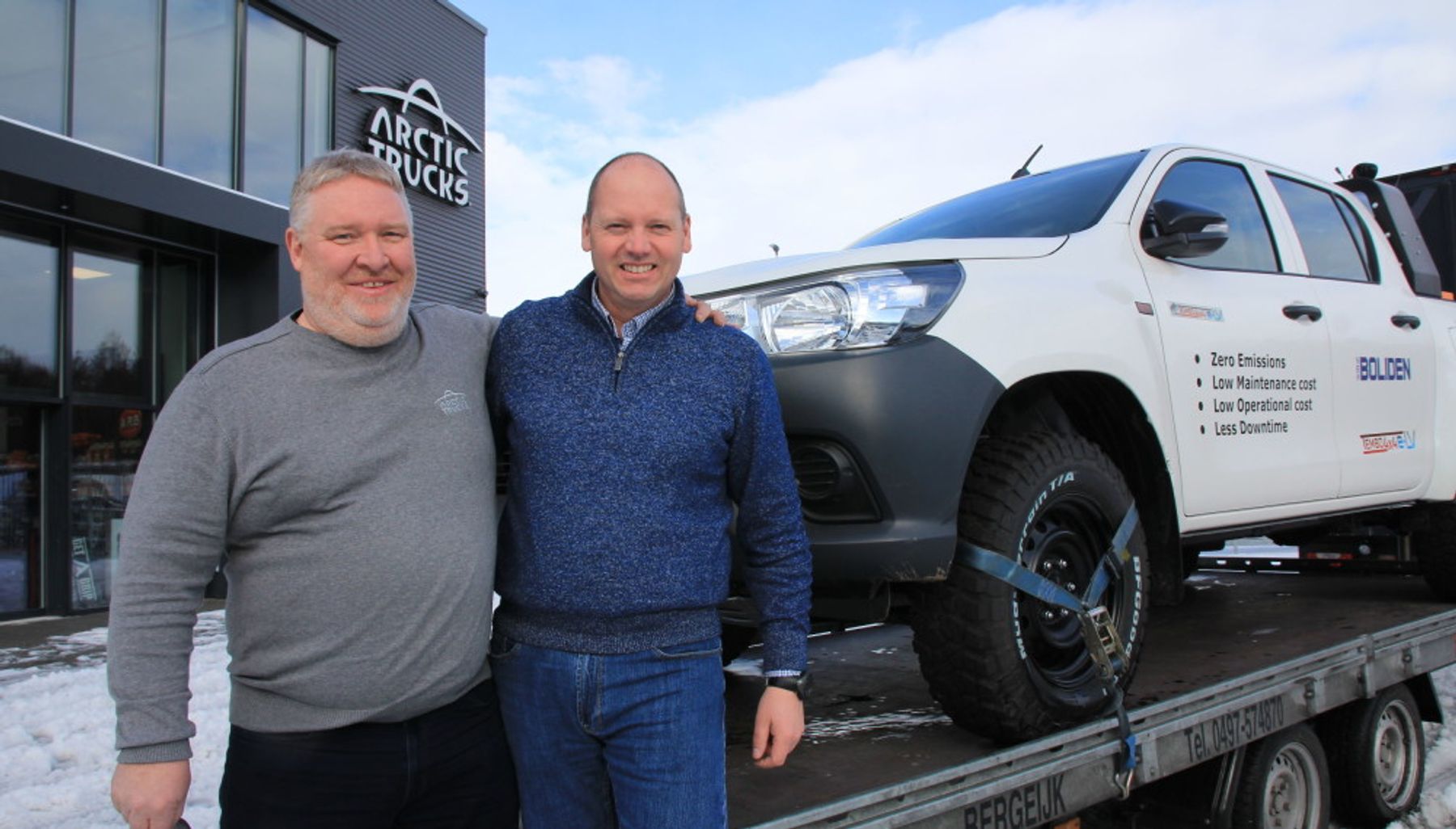 Two men smile outside an Arctic Trucks building beside a Boliden-branded electric Hilux on a trailer