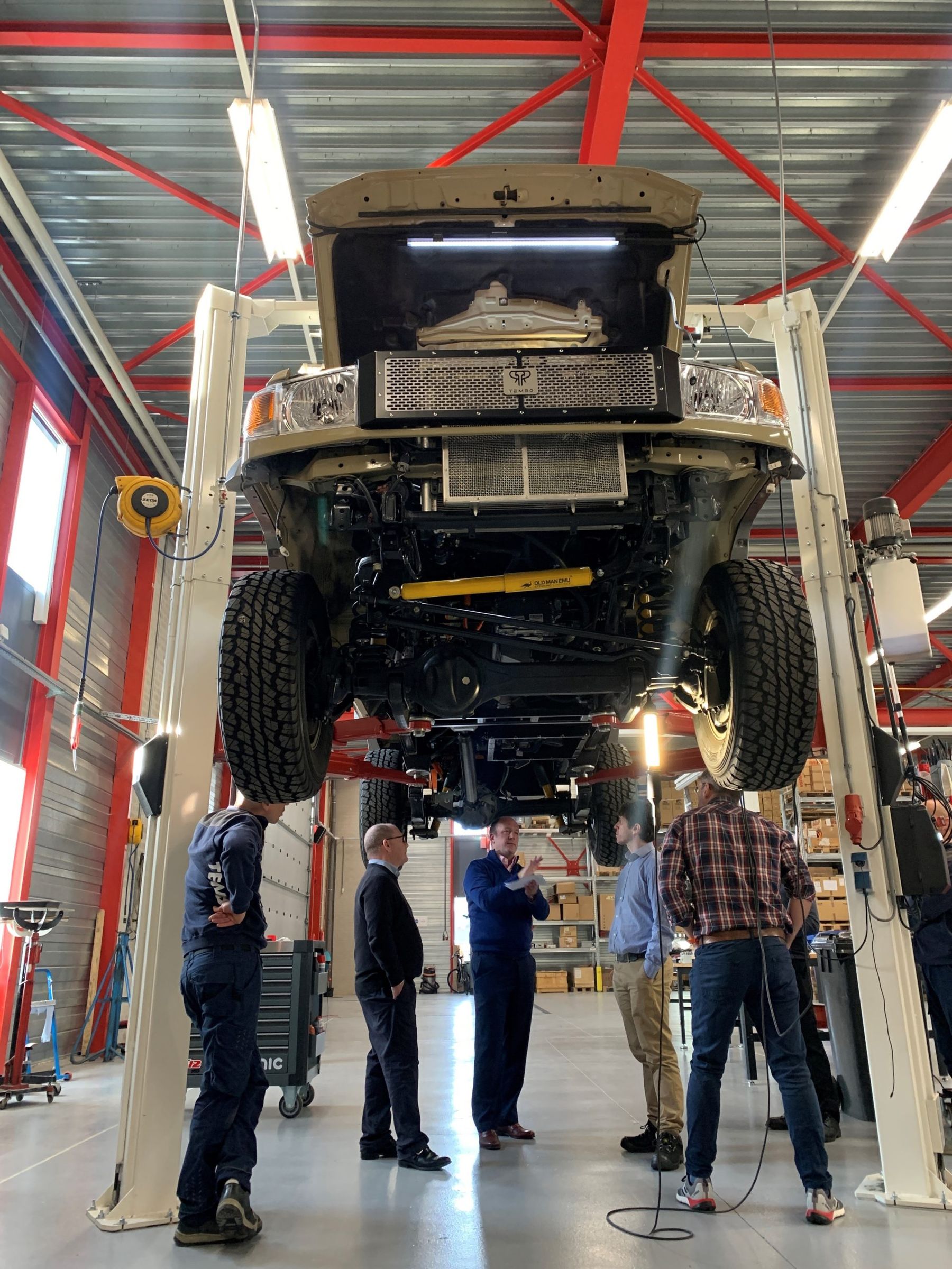 Visitors standing beneath an electric Land Cruiser 70 raised on a vehicle lift in Tembo’s Eindhoven workshop