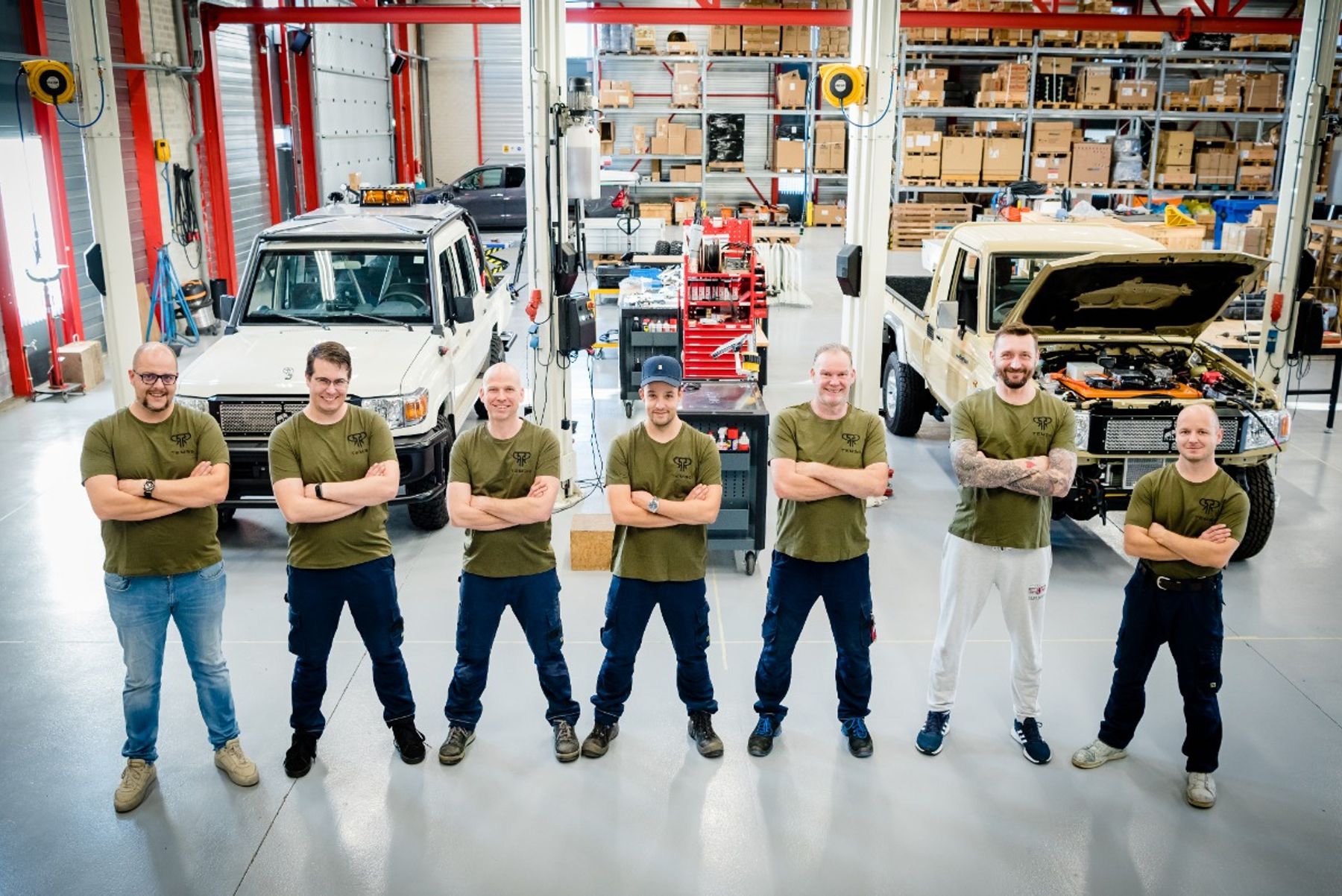 Seven Tembo team members in branded T-shirts, arms crossed, in the workshop with two Land Cruiser 70 vehicles behind