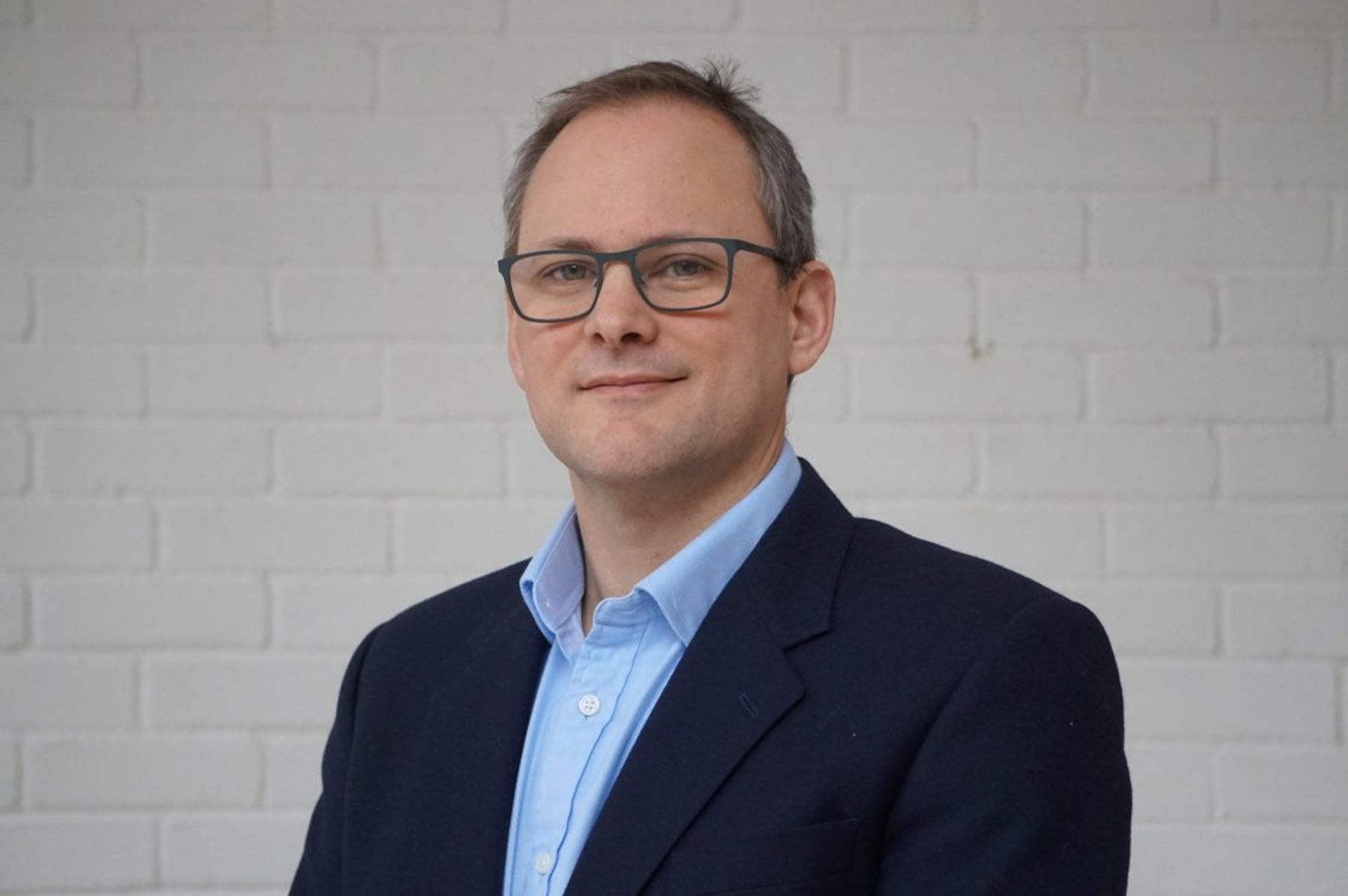 Portrait of James Howell-Richardson, VivoPower General Counsel, in a navy blazer against a white brick wall