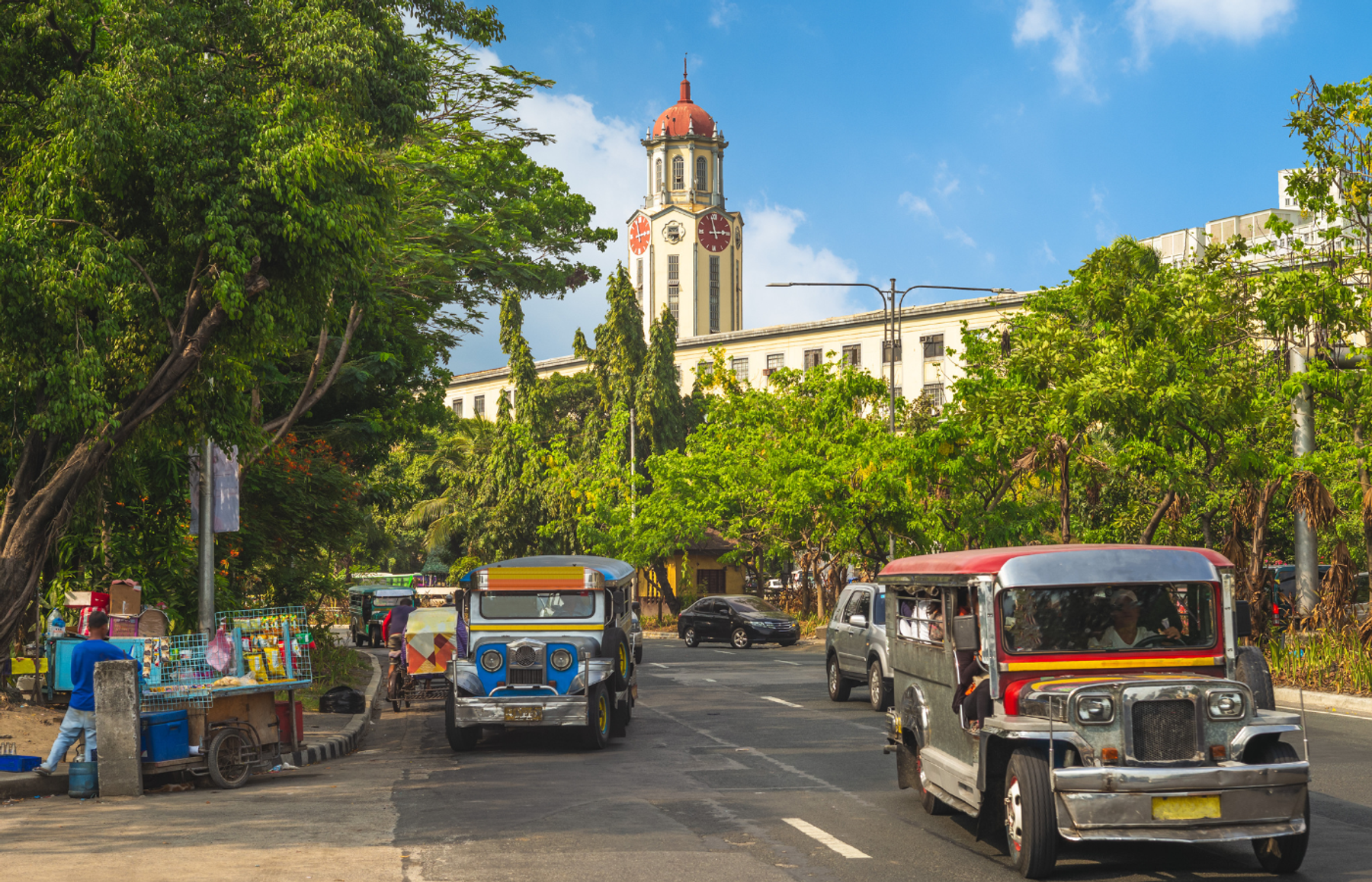 Traditional jeepneys driving along a tree-lined city street in the Philippines, with a clock tower building behind