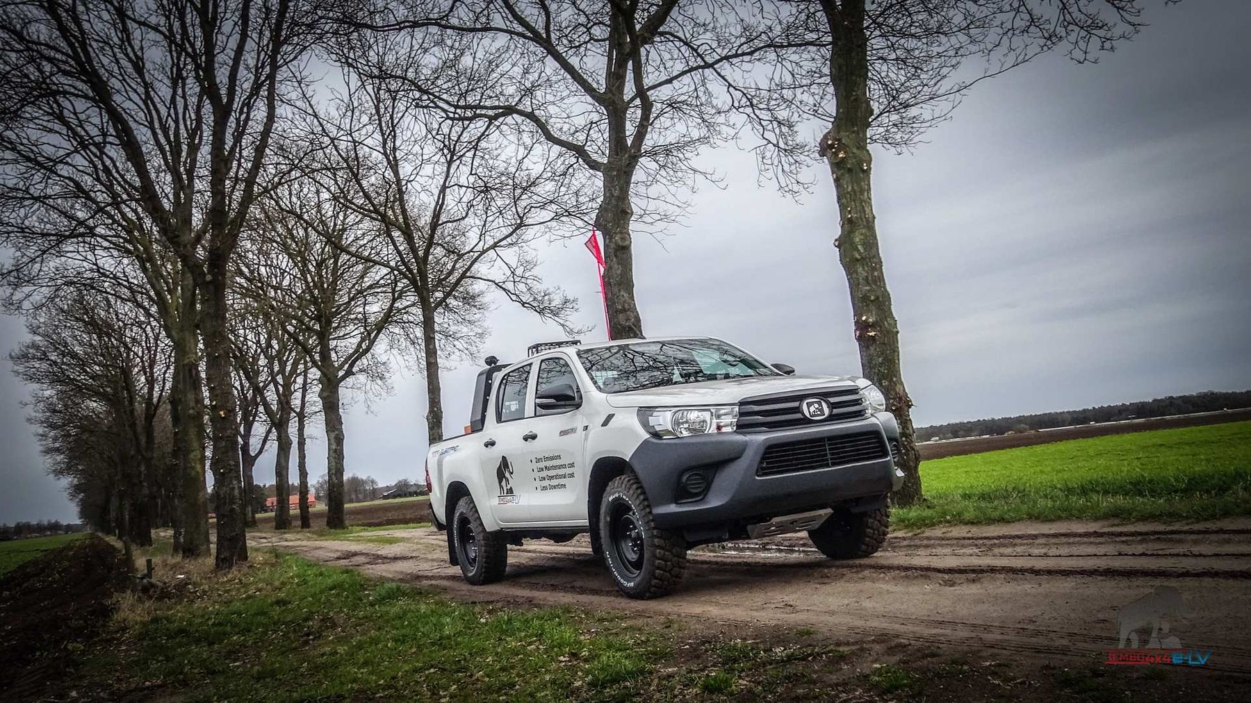 Electric Toyota Hilux dual-cab conversion on a dirt track lined with bare trees beside green farmland