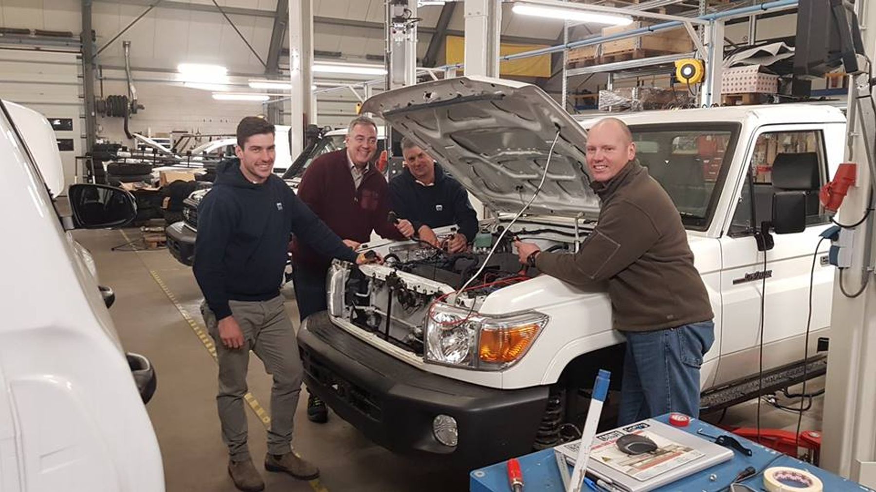Four men working on the wiring under the open bonnet of a white Toyota Land Cruiser 70 series in a workshop