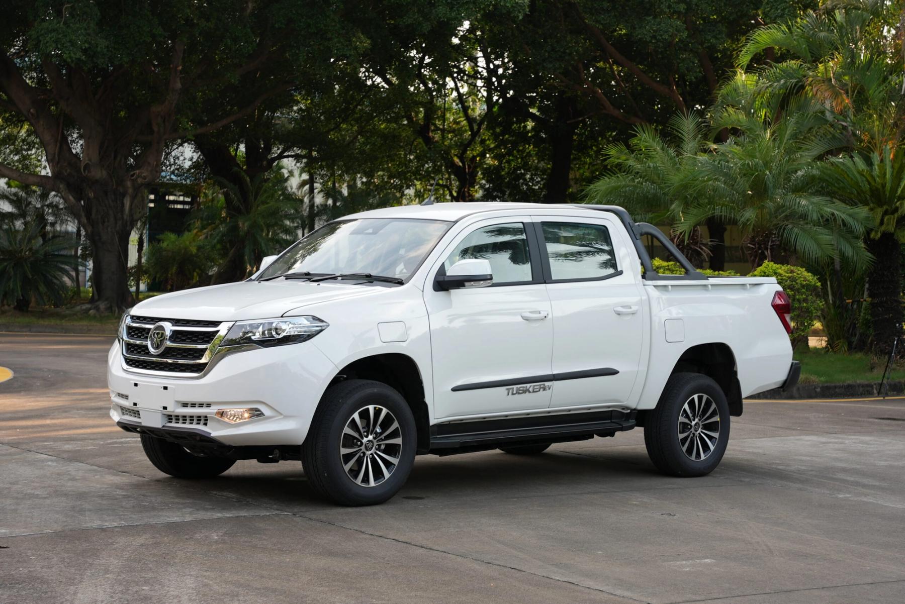 White Tembo Tusker dual-cab electric ute with sports bar parked on a forecourt in front of tropical trees