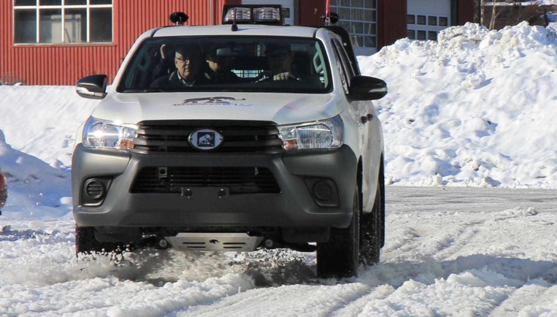 White electric Toyota Hilux with Tembo elephant badge driving through snow past a red building