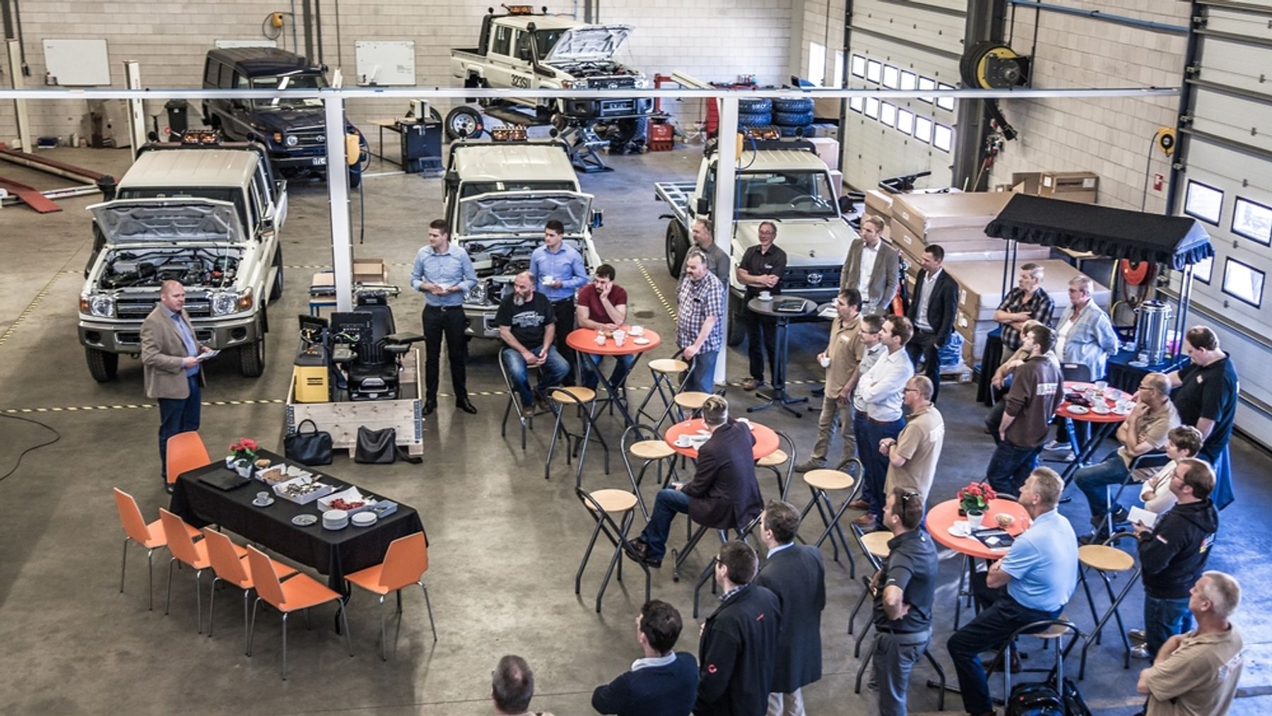 Overhead view of guests at the Tembo 4x4 e-LV prototype launch in the workshop, among Land Cruisers with bonnets open