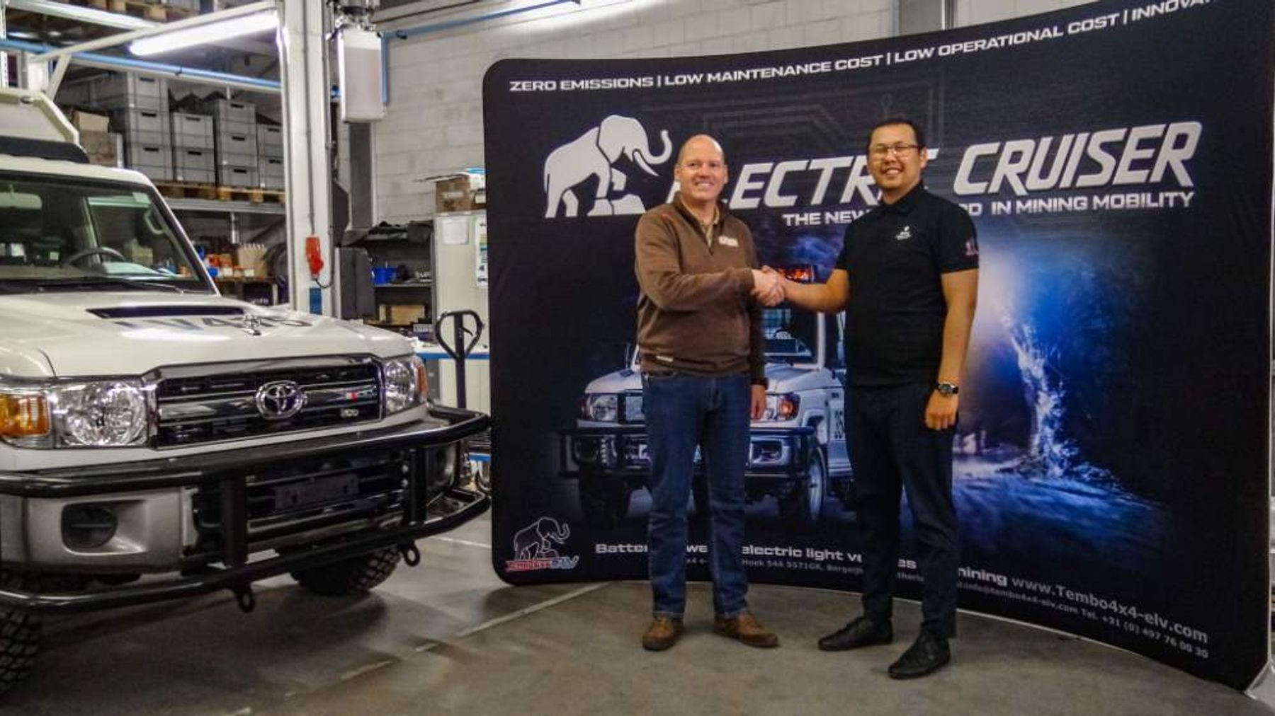 Two men shake hands in front of an Electric Cruiser backdrop in a workshop, beside a white Toyota Land Cruiser