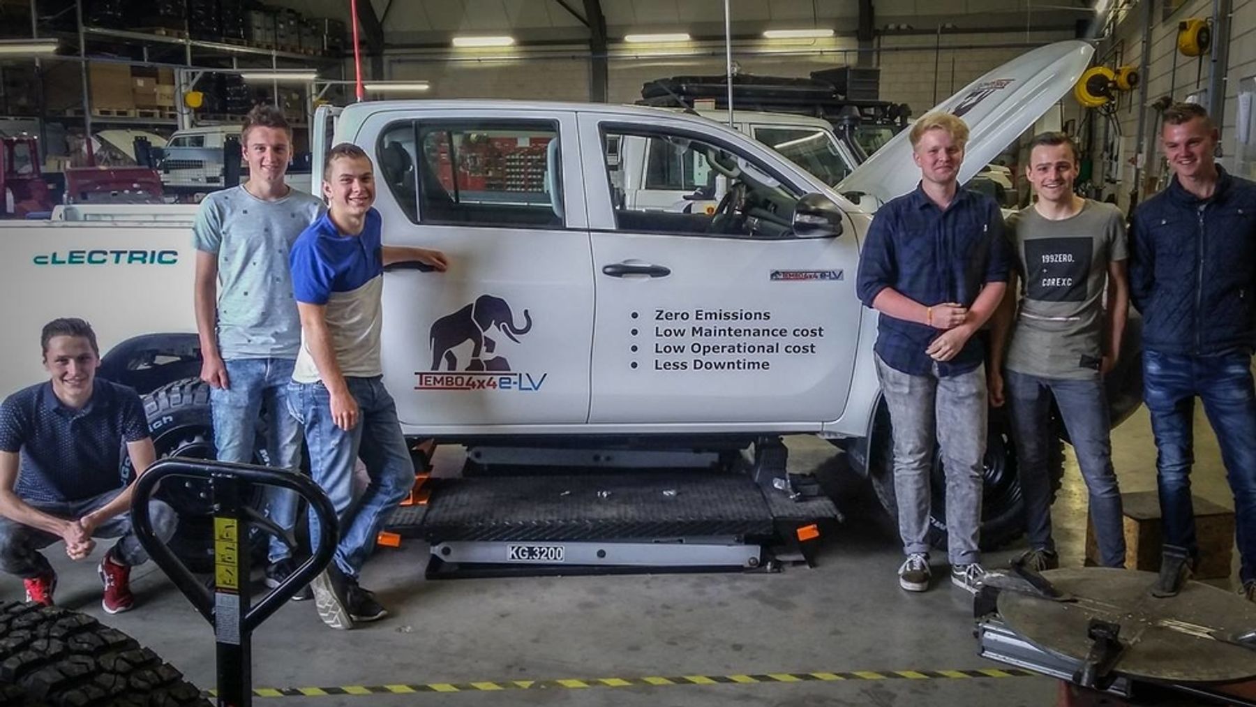 Six students posing beside a Tembo 4x4 e-LV dual-cab ute on a vehicle lift in a workshop on student trial day