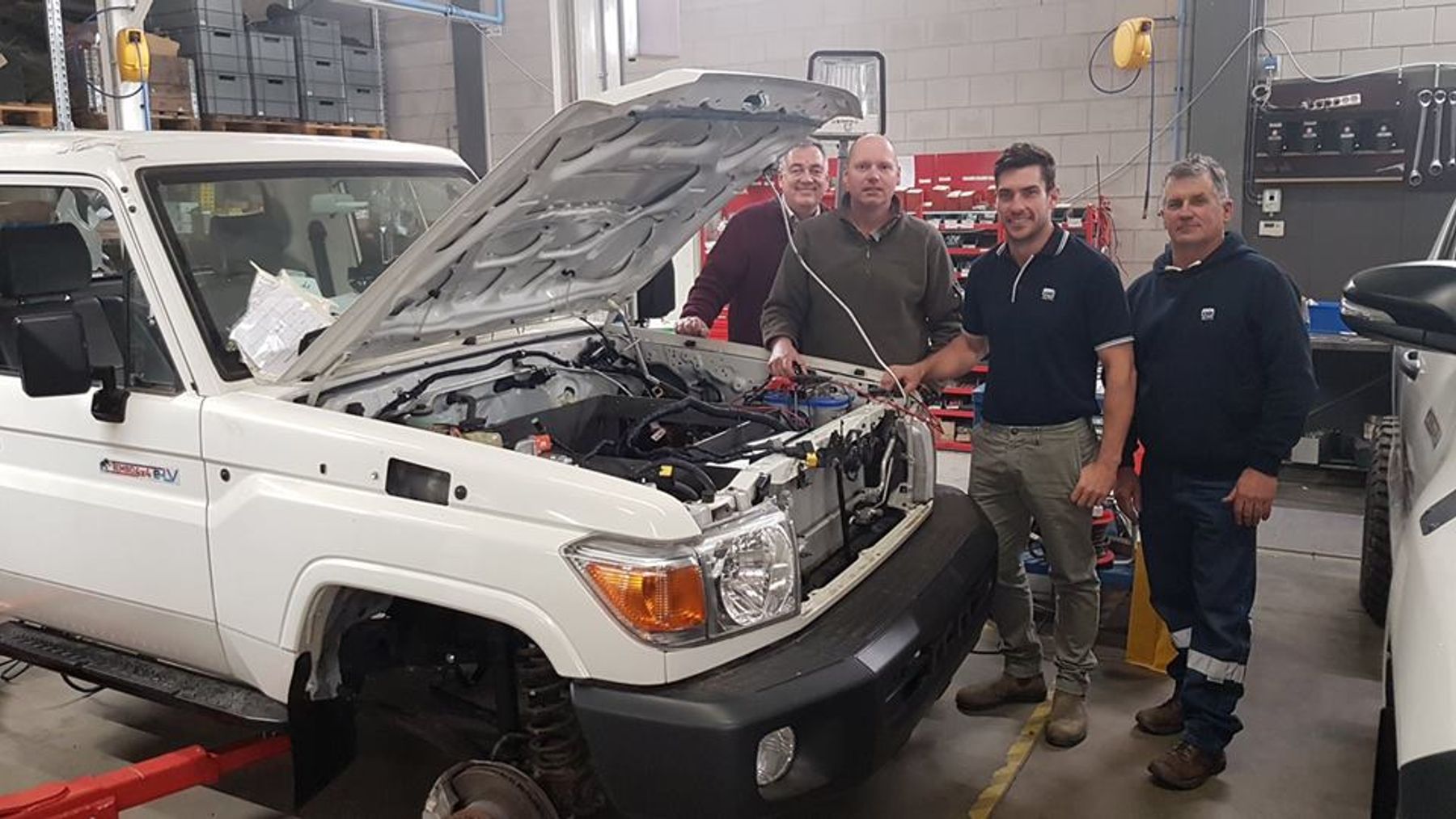 Four men beside a white Toyota Land Cruiser 70 series with its bonnet open, showing new wiring in a workshop