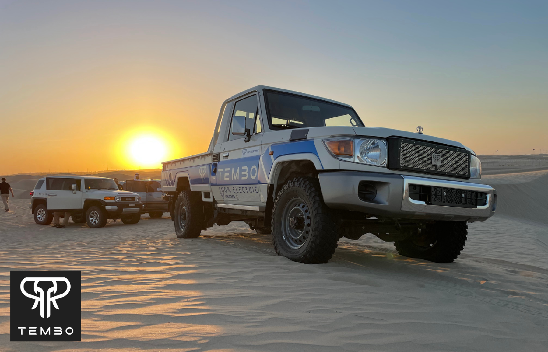 Tembo-liveried 100% electric Land Cruiser 70 series pickup on desert sand at sunset, Toyota FJ Cruiser and people behind