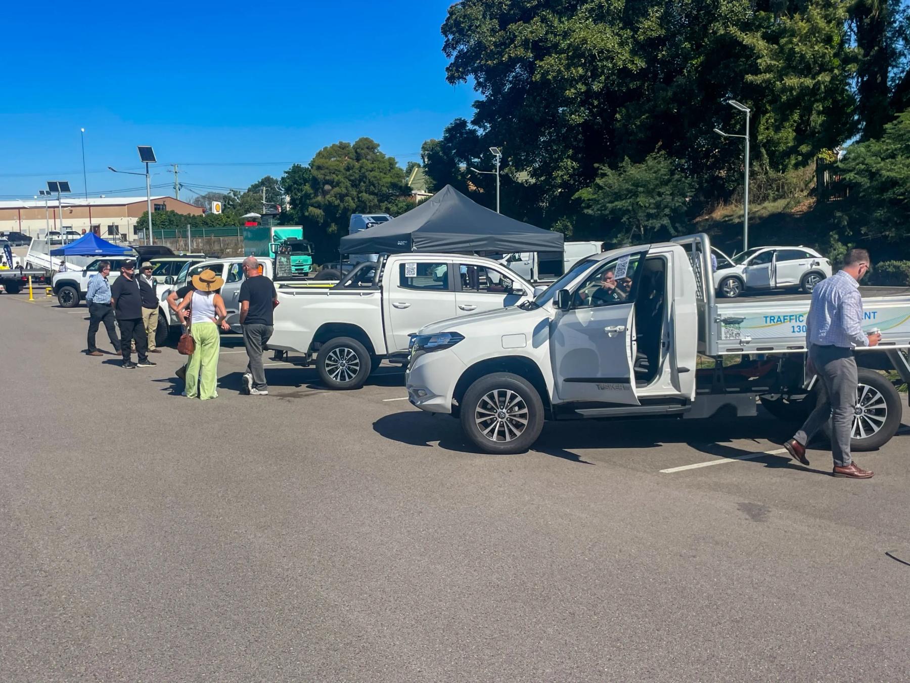 White Tembo Tusker single-cab electric ute with alloy tray on display in a car park as visitors view other electric vehicles