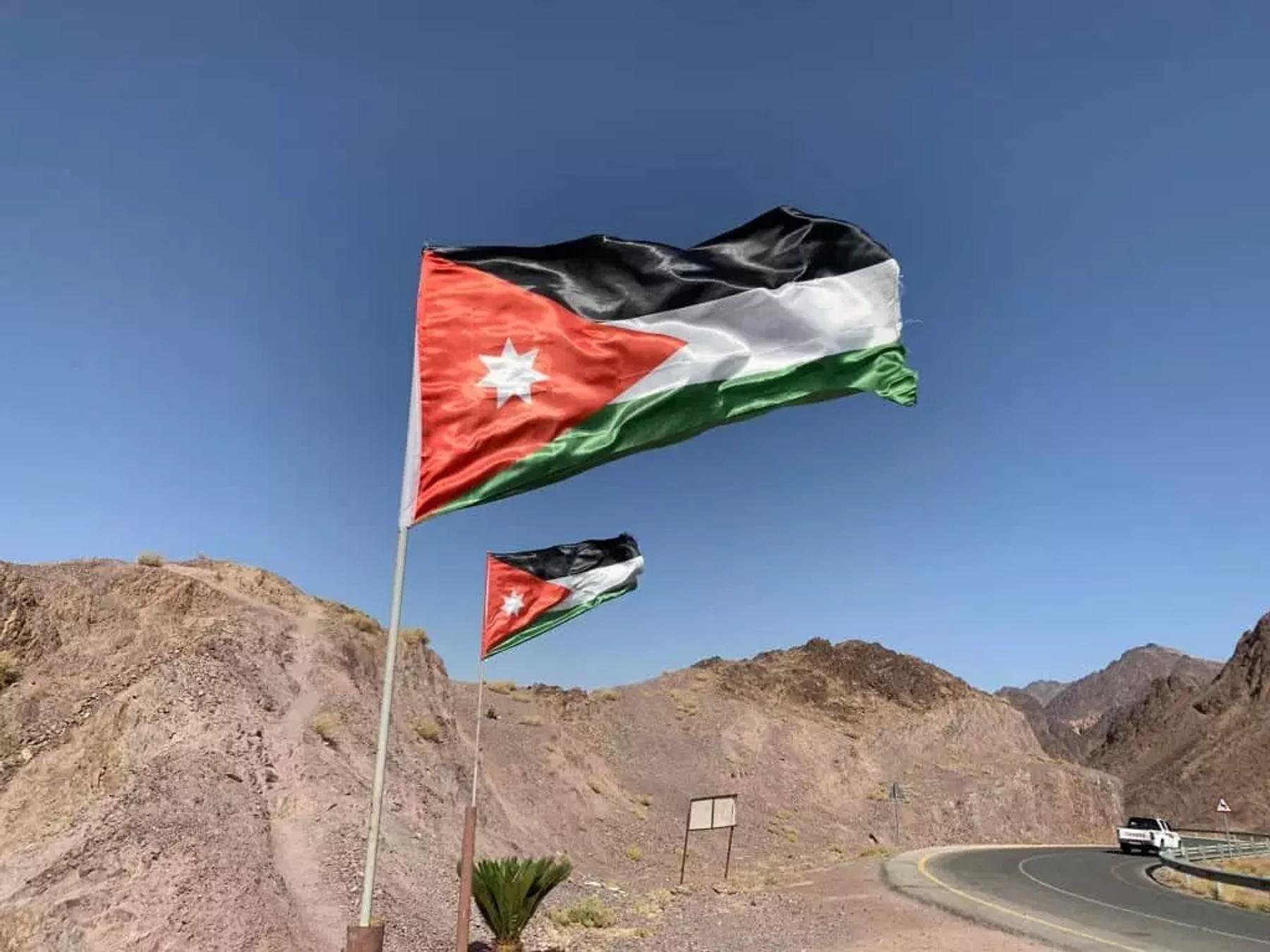 Jordanian flags flying beside a winding road through rocky desert hills, with a white pick-up in the distance