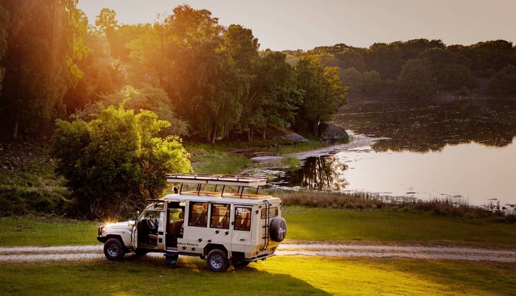 Land Cruiser safari vehicle with open sides parked on a grassy track beside a lake at sunset