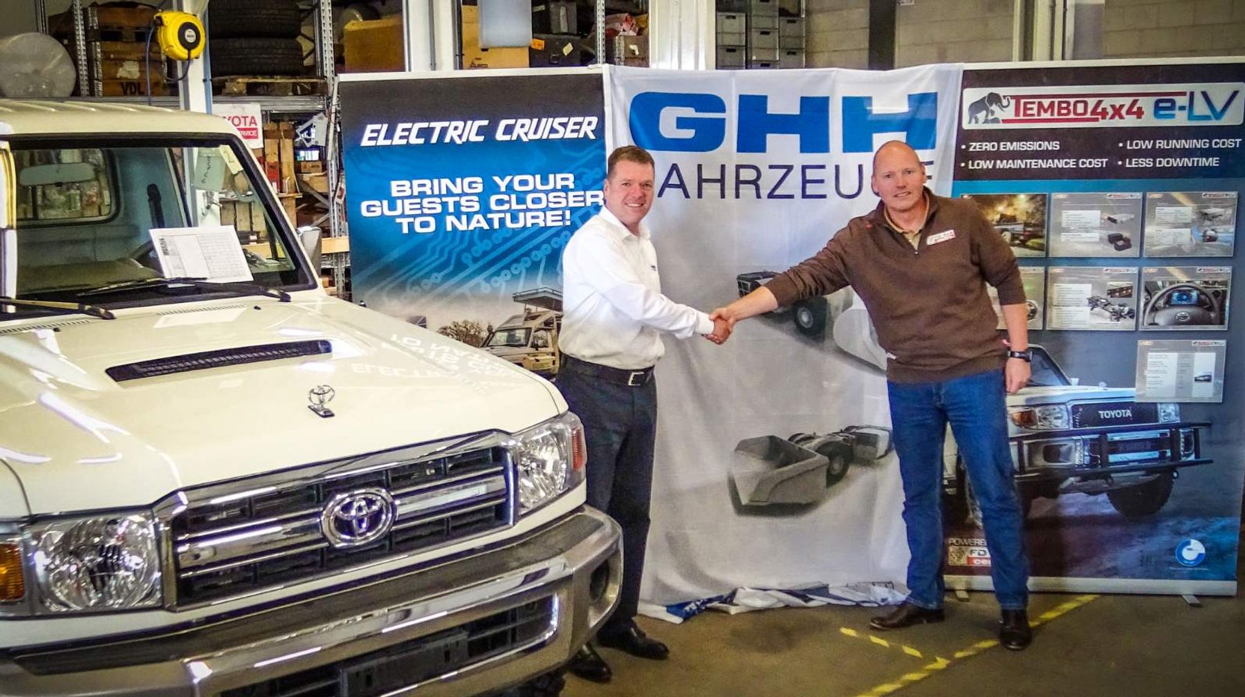 Handshake in front of GHH Fahrzeuge and Tembo 4x4 e-LV banners beside a white Toyota Land Cruiser in a workshop