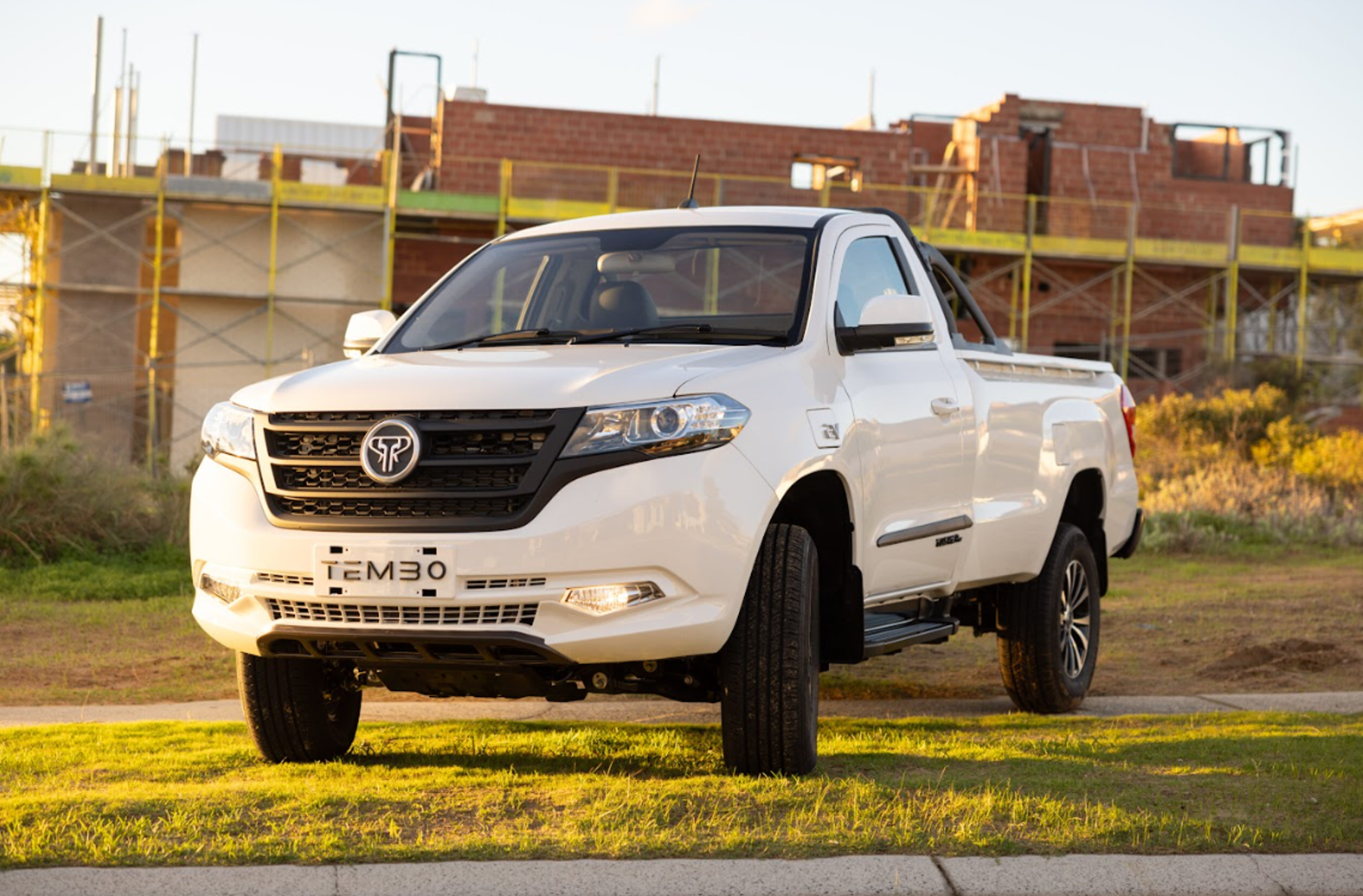 White Tembo Tusker single-cab electric ute parked on grass in front of a brick house under construction