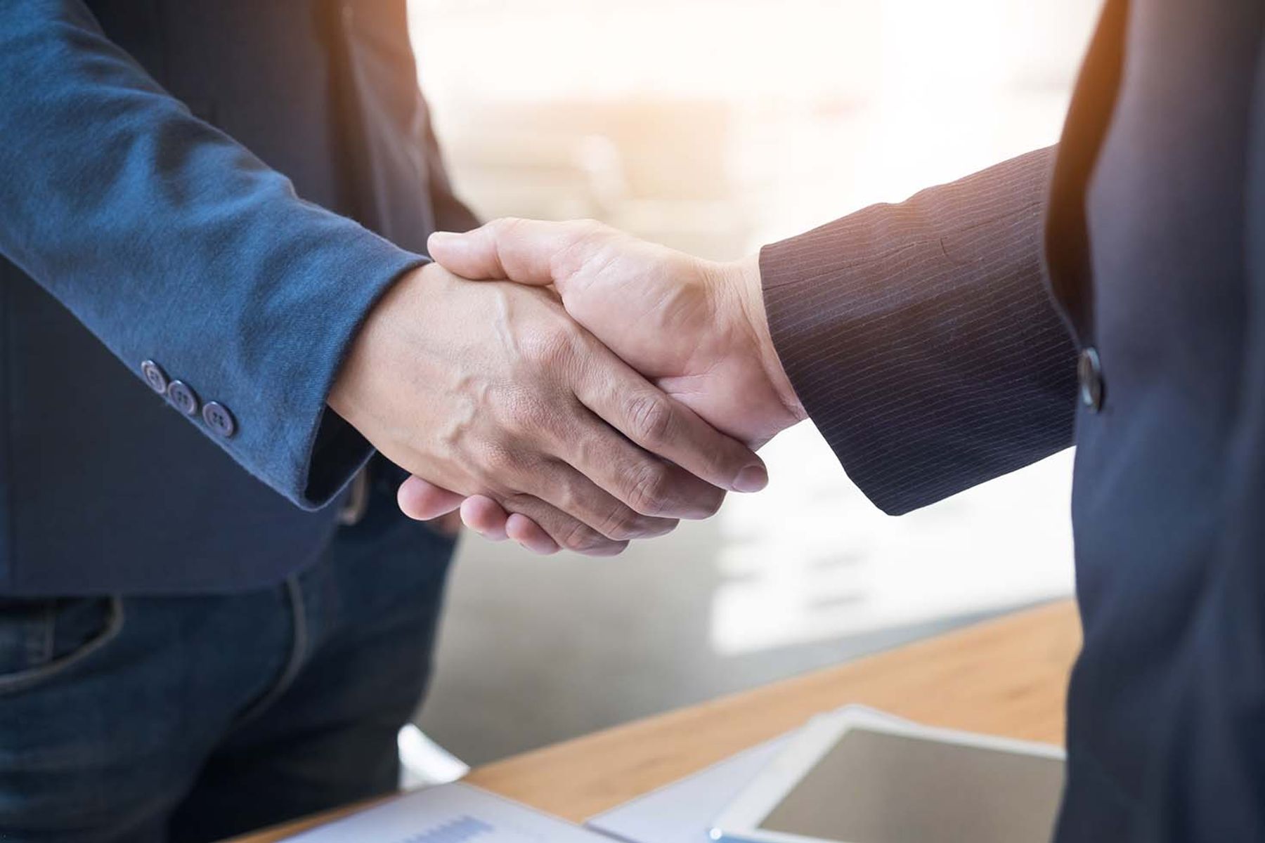 Two business people in suits shaking hands over a desk with documents and a tablet