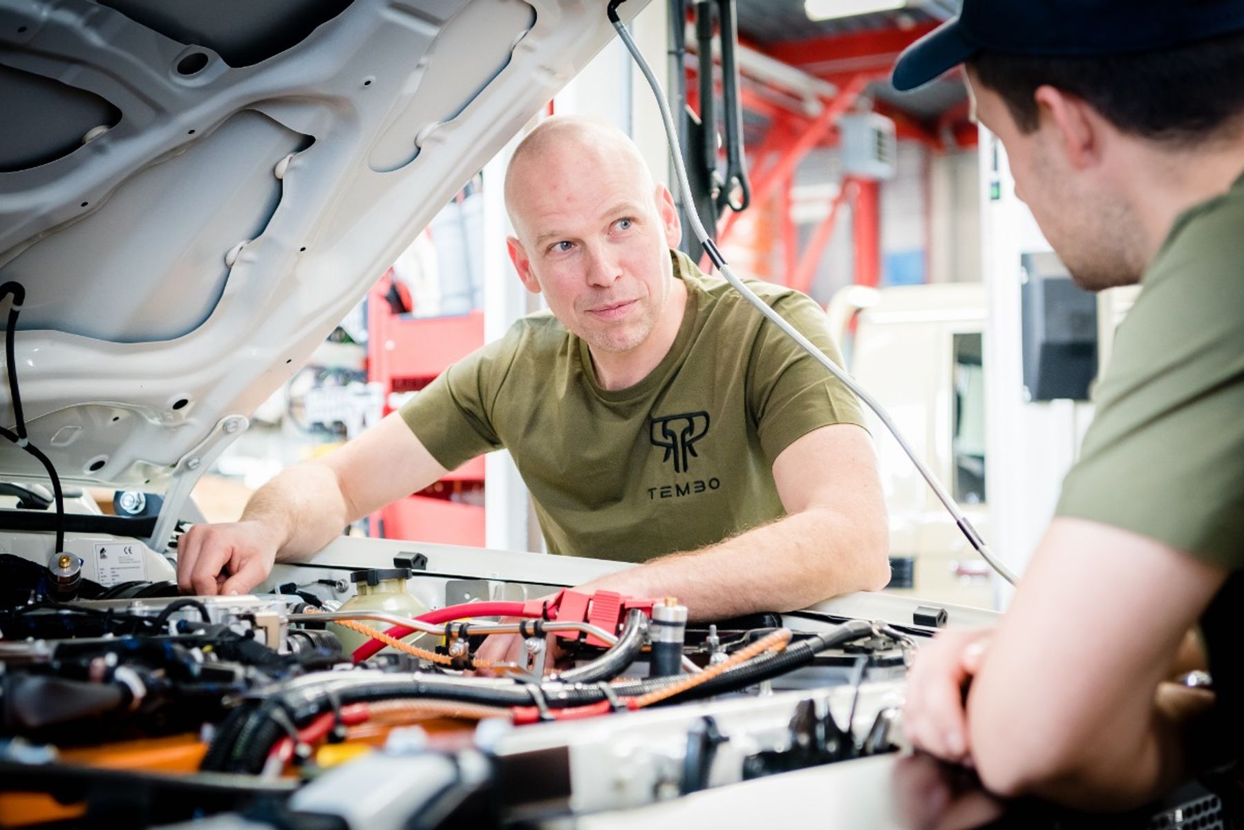 Tembo technicians in branded T-shirts talk over the open bonnet of a vehicle with electric drivetrain wiring