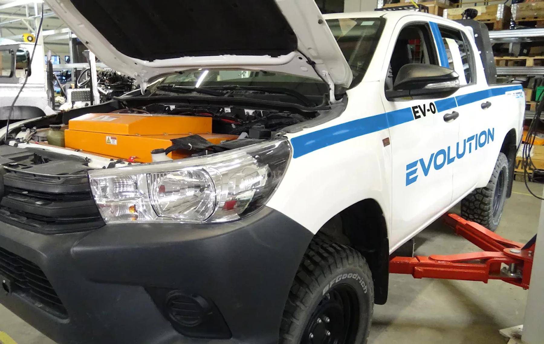 Evolution-branded Toyota Hilux on a workshop hoist with bonnet open, showing an orange electric drive unit