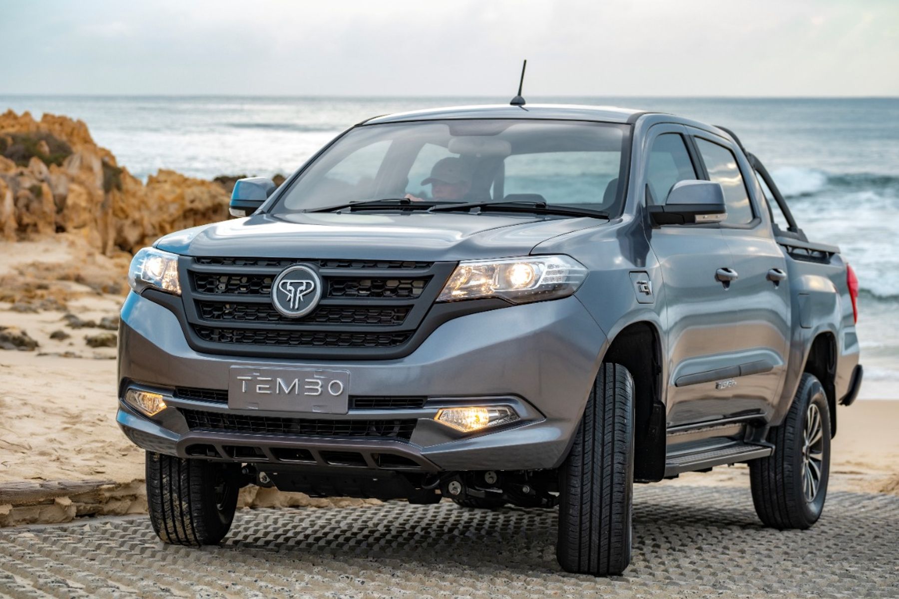 Grey Tembo Tusker dual-cab electric ute on beach matting beside rocks and the ocean, driver at the wheel
