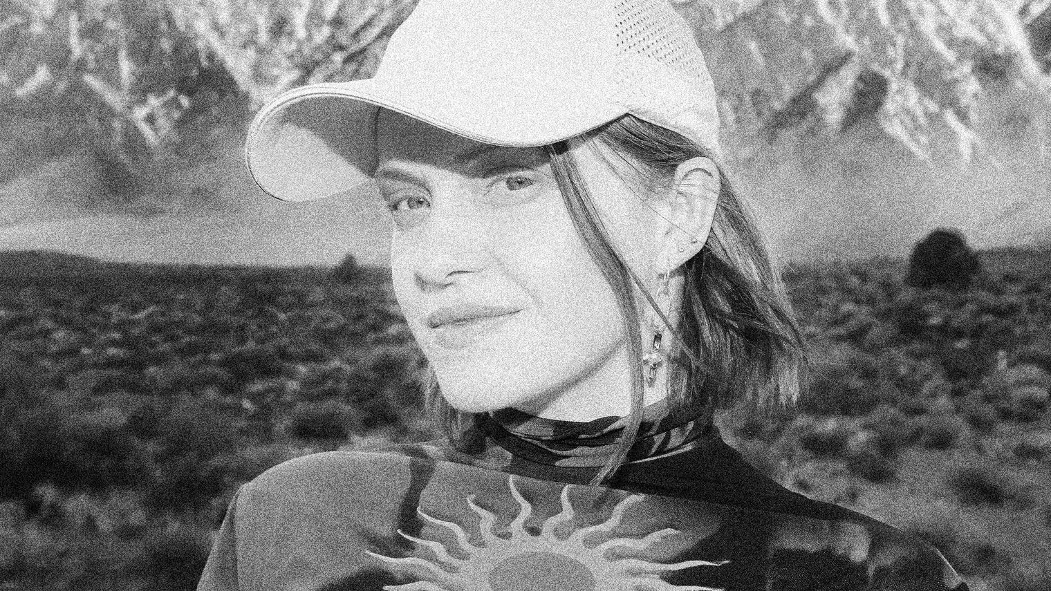 Greyscale portrait of Kučka wearing a cap and ornate earrings, in the background grass plains and distant mountains can be seen.