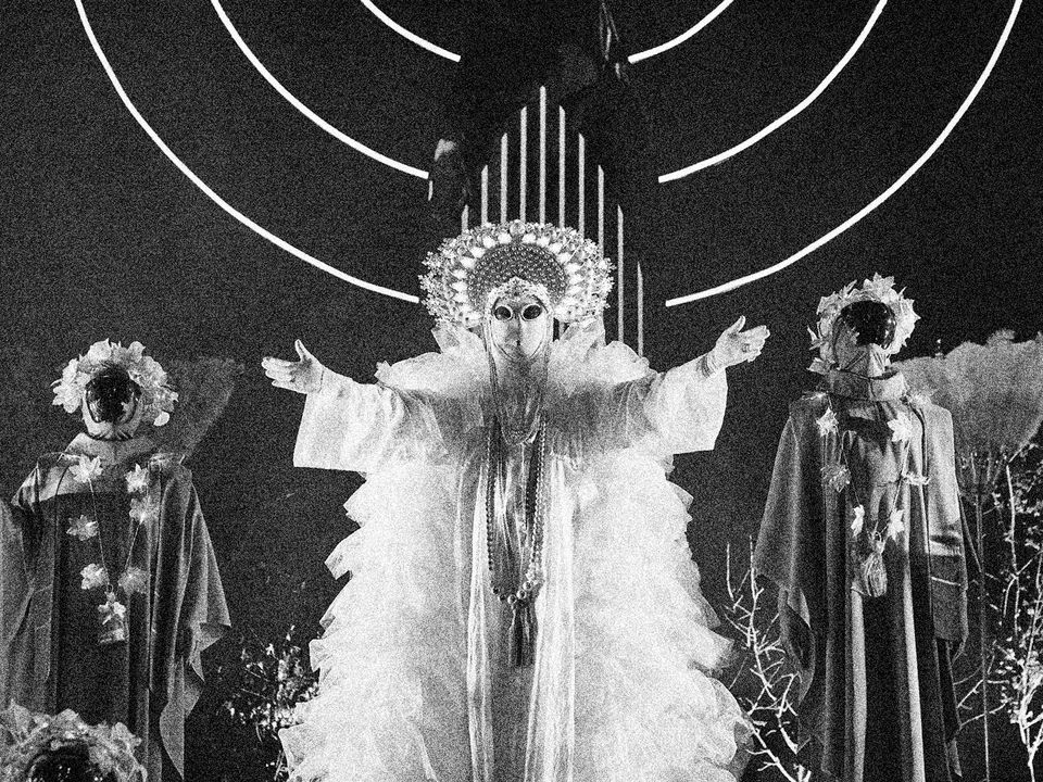 Pope Alice garbed in an extravagant outfit stands with arms outstretched. 