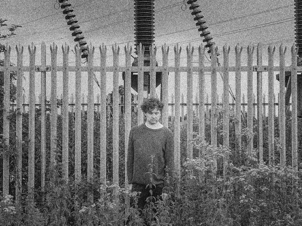 Nathan Fake stands in-front of a spiked metal fence, behind that is electrical infrastructure. 