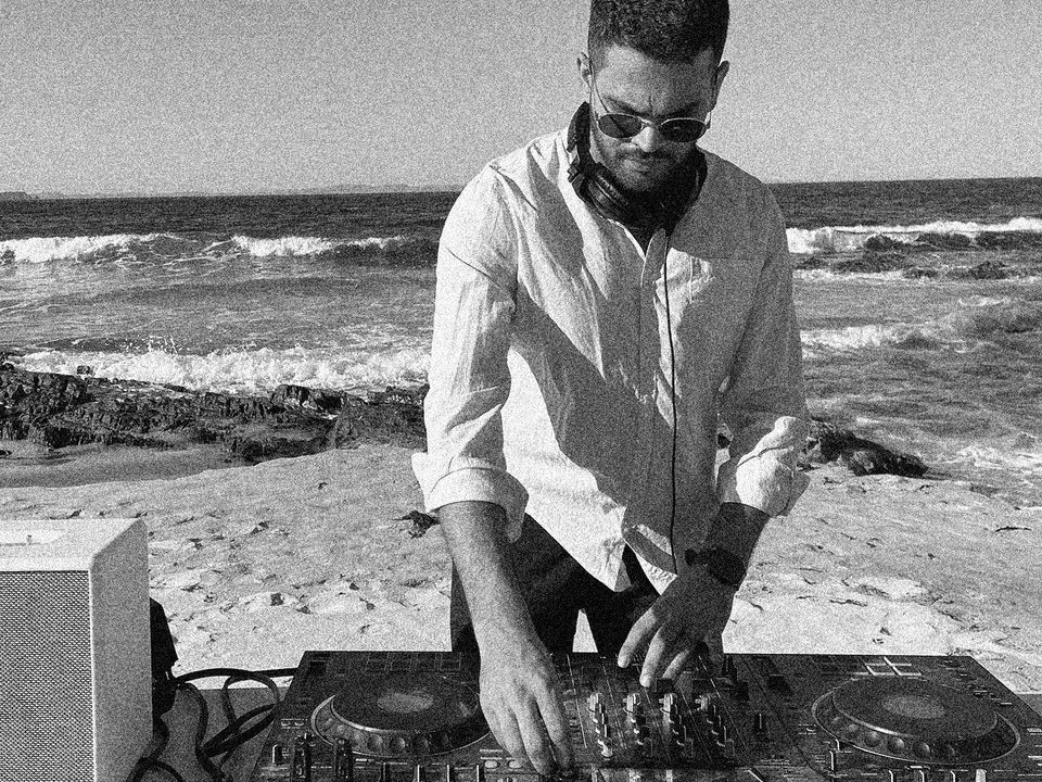 DHAIRYA adjusts knobs on a DJ deck, in the background are waves at a beach.