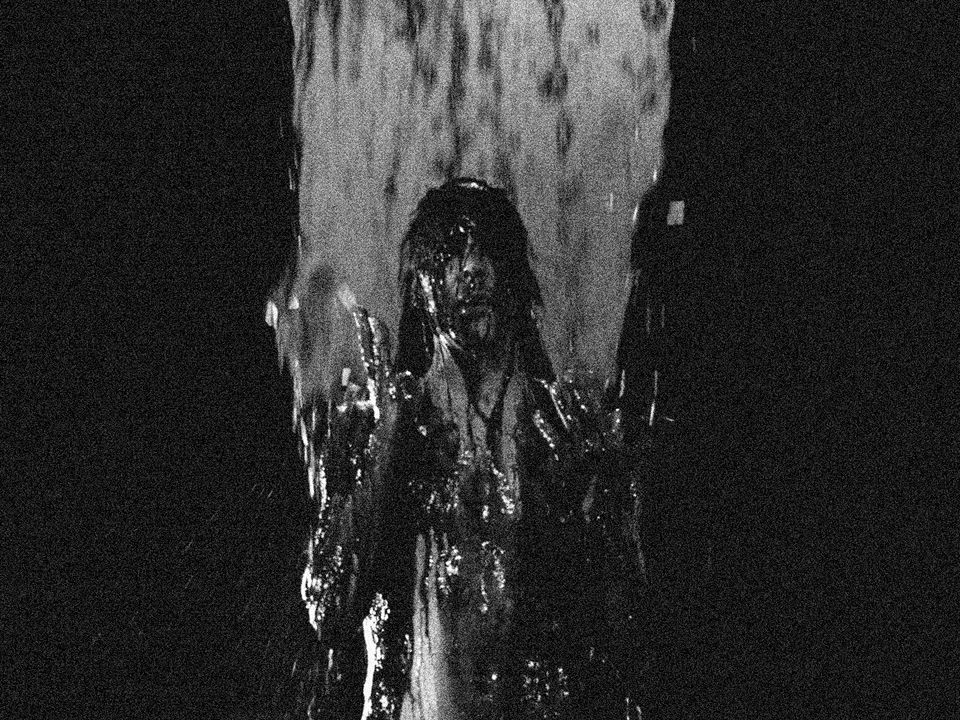 A shirt-less man stands covered in dark liquid, looking upwards as more falls from above.