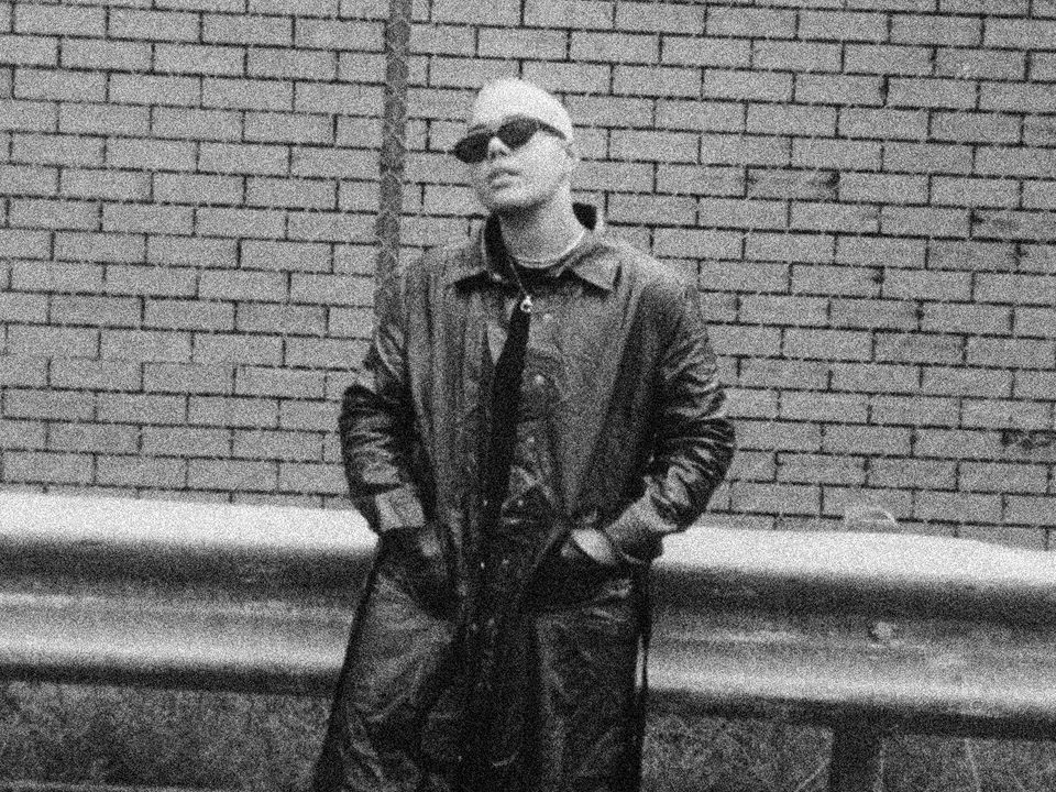 Jesswar poses against a barrier wearing a trench-coat and dark sunglasses.