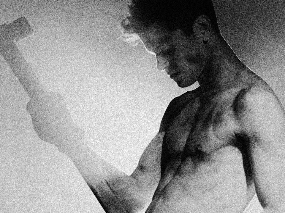 Greyscale image of a shirt-less Perfume Genius holding a sledgehammer in one hand, lifted to head height.
