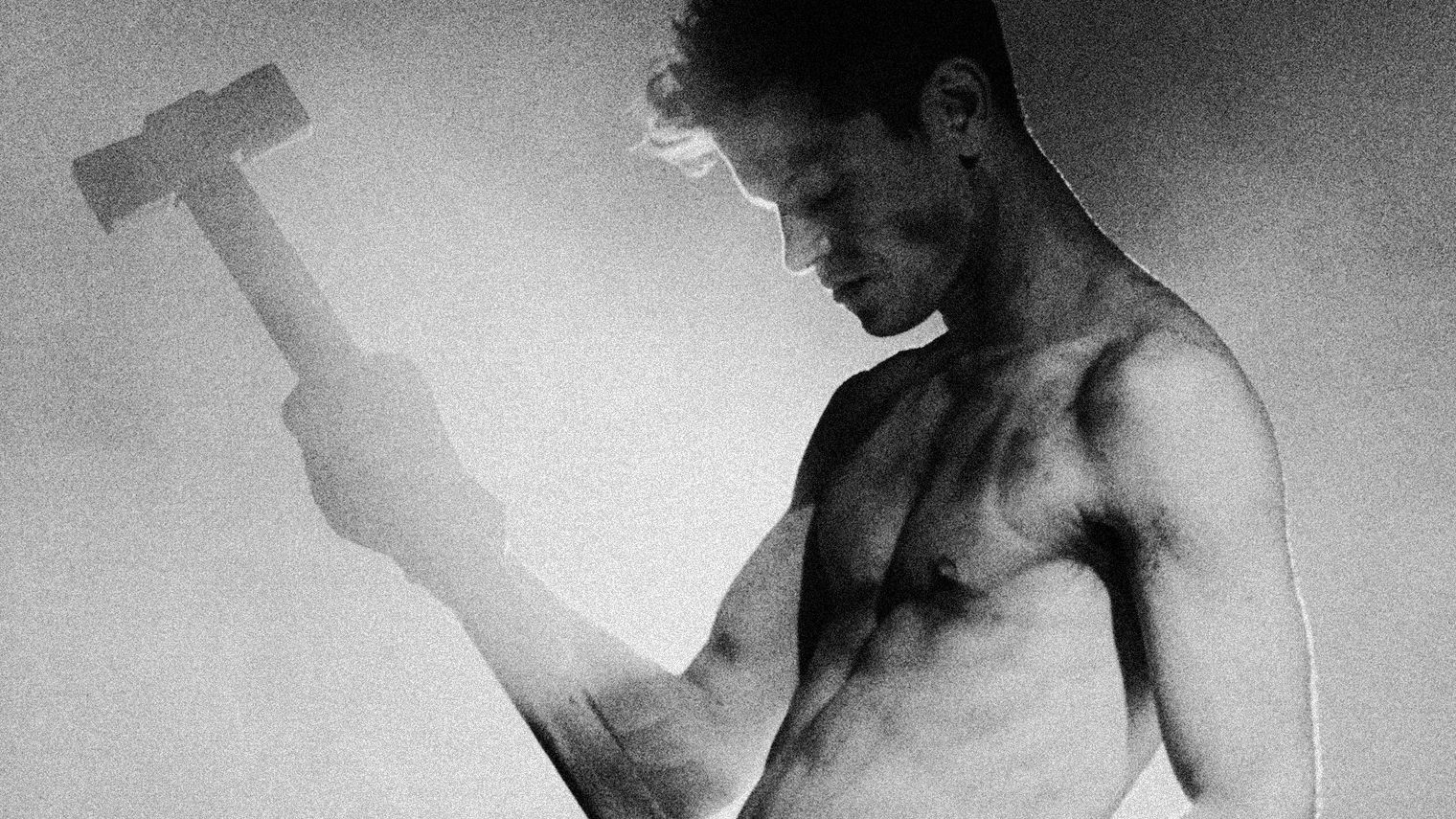 Greyscale image of a shirt-less Perfume Genius holding a sledgehammer in one hand, lifted to head height.