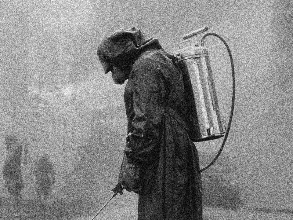 A person wearing a hazmat suit uses a hose which is attached to a metal canister on their back.