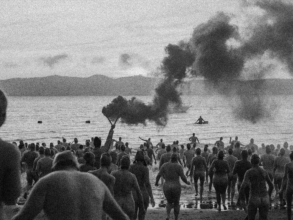 Pictured from behind, hundreds of nude swimmers run into the ocean, one person holds a smoking beacon above their head.