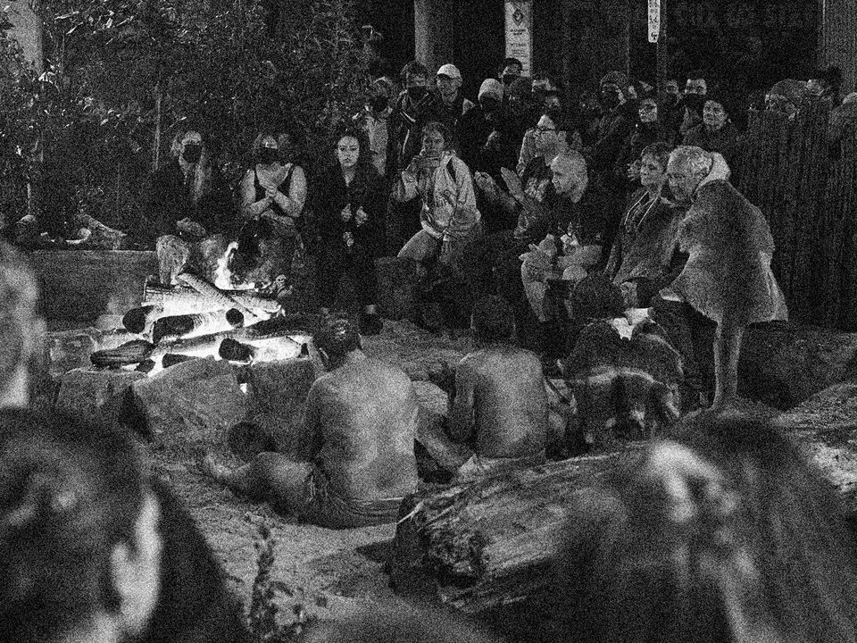 A large group of people sit around a burning outdoor fireplace.