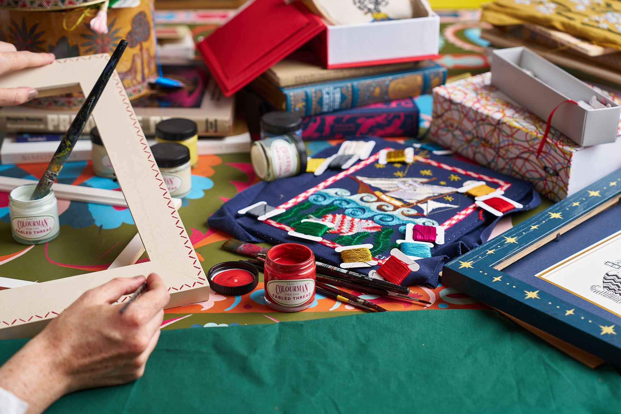 Picture frame being hand painted on a table surrounded by embroidery threads and art