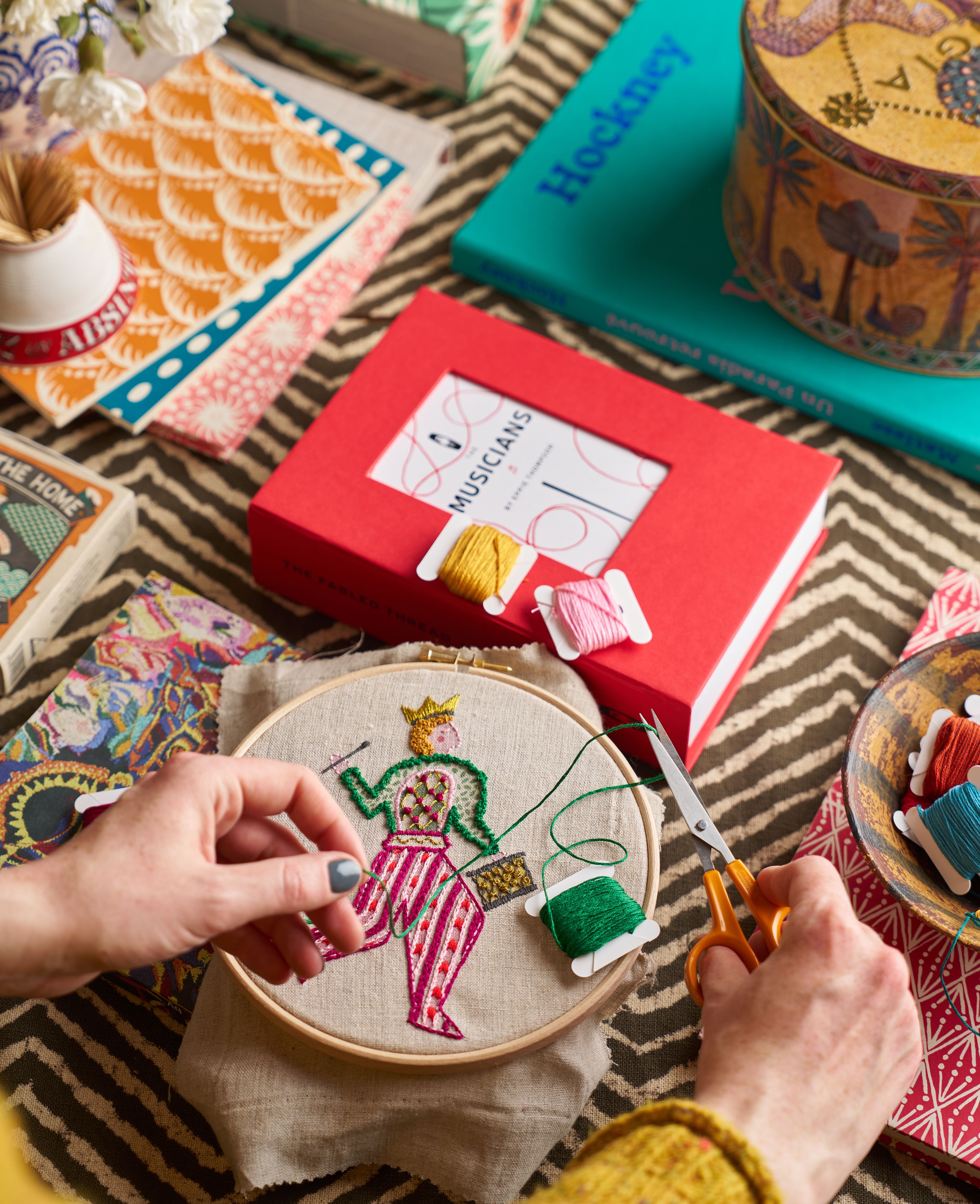 Embroidery kit of the Drummer on a table with books, tins and embroidery thread